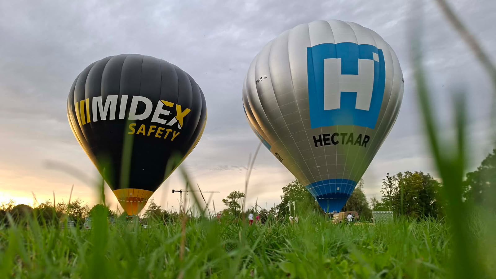 Twee hete luchtballonnen met logo's van Midex Safety en Hectaar opstijgend boven een grasveld bij zonsopgang