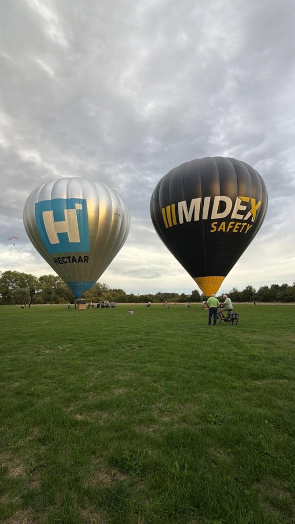 Twee hete luchtballonnen, zilver met blauw logo Hectaar en zwart met geel Midex Safety, stijgen op vanaf groen veld met wolkenlucht