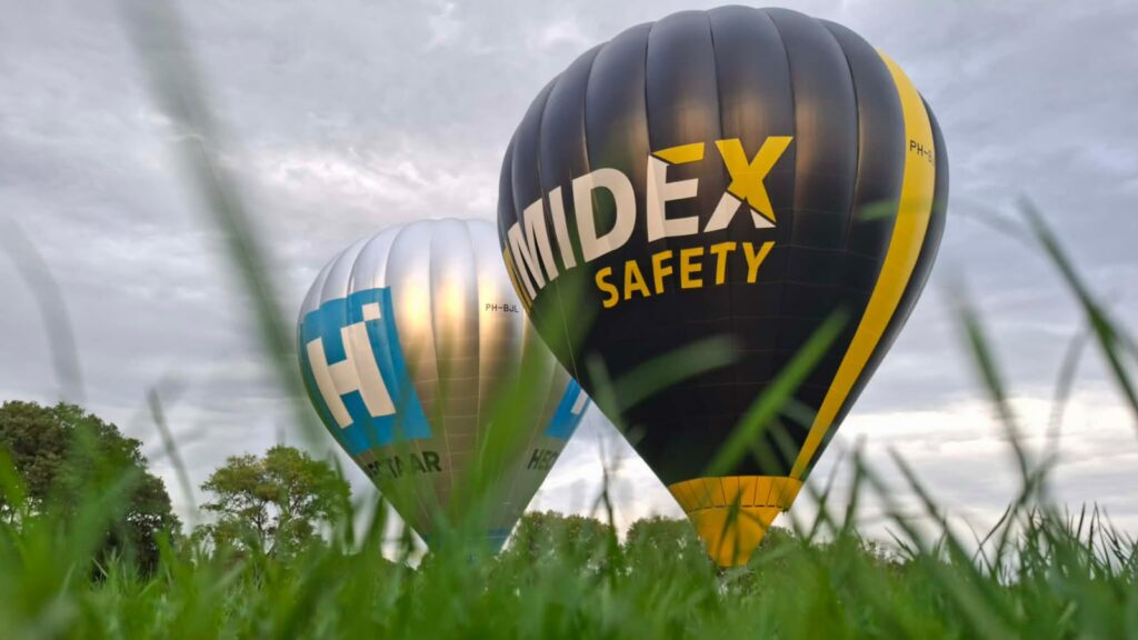 Twee kleurrijke hot-air balloons met bedrijfslogo's zweven laag boven grasland onder een bewolkte hemel